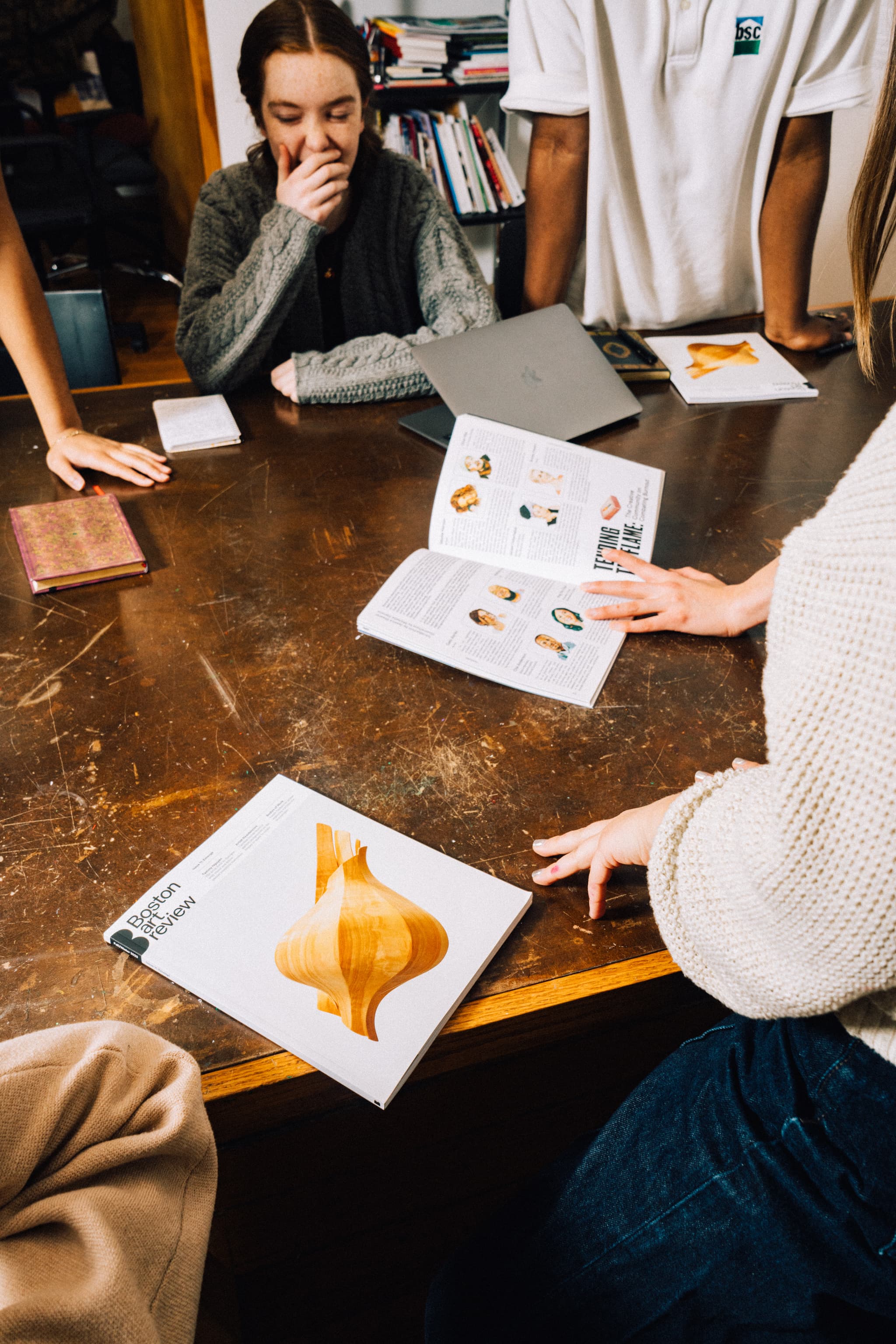 Dylan, a young light-skinned woman with red hair, sits at the table looking towards an open copy of Issue 11.