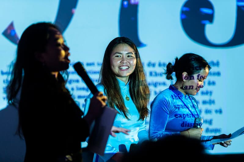 Three women stand in front of a screen projecting words, the middle figure smiling.